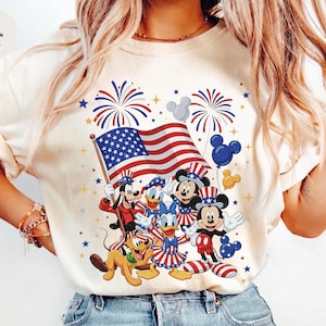 May include: Ivory-colored t-shirt featuring a patriotic design with Mickey Mouse, Minnie Mouse, Donald Duck, and Goofy. The design includes the American flag, fireworks, and Mickey Mouse-shaped balloons. The text "Colors Ivory" is visible.