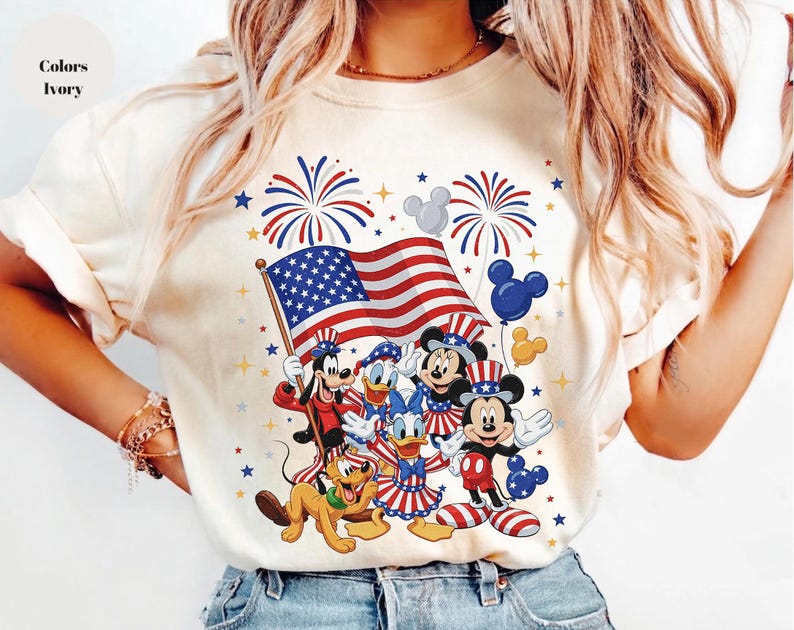 May include: Ivory-colored t-shirt featuring a patriotic design with Mickey Mouse, Minnie Mouse, Donald Duck, and Goofy. The design includes the American flag, fireworks, and Mickey Mouse-shaped balloons. The text "Colors Ivory" is visible.