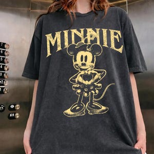 May include: Dark gray t-shirt featuring a yellow graphic of Minnie Mouse and the word "MINNIE" in a vintage font. The graphic shows Minnie Mouse with a heart-shaped design. The shirt has short sleeves and a relaxed fit.
