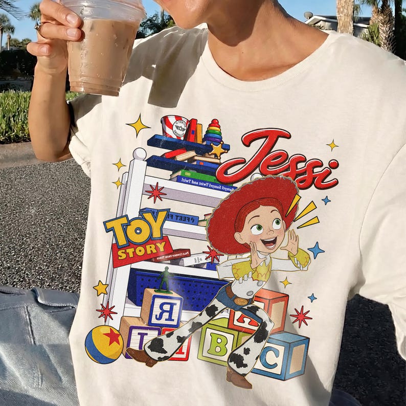 Puede incluir: Camiseta color crema con un gr&aacute;fico del personaje Jessie de Toy Story. El dise&ntilde;o incluye el texto "Toy Story" y "Jessie" en una fuente l&uacute;dica, junto con estrellas coloridas y bloques de juguetes.