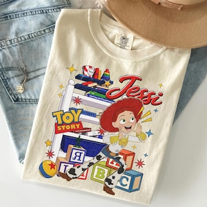 Puede incluir: Camiseta color crema con el personaje Jessi de Toy Story. El dise&ntilde;o incluye las palabras "Toy Story" y "Jessi" en rojo, junto con bloques de juguetes coloridos y estrellas. La camiseta est&aacute; doblada sobre unos vaqueros azules.