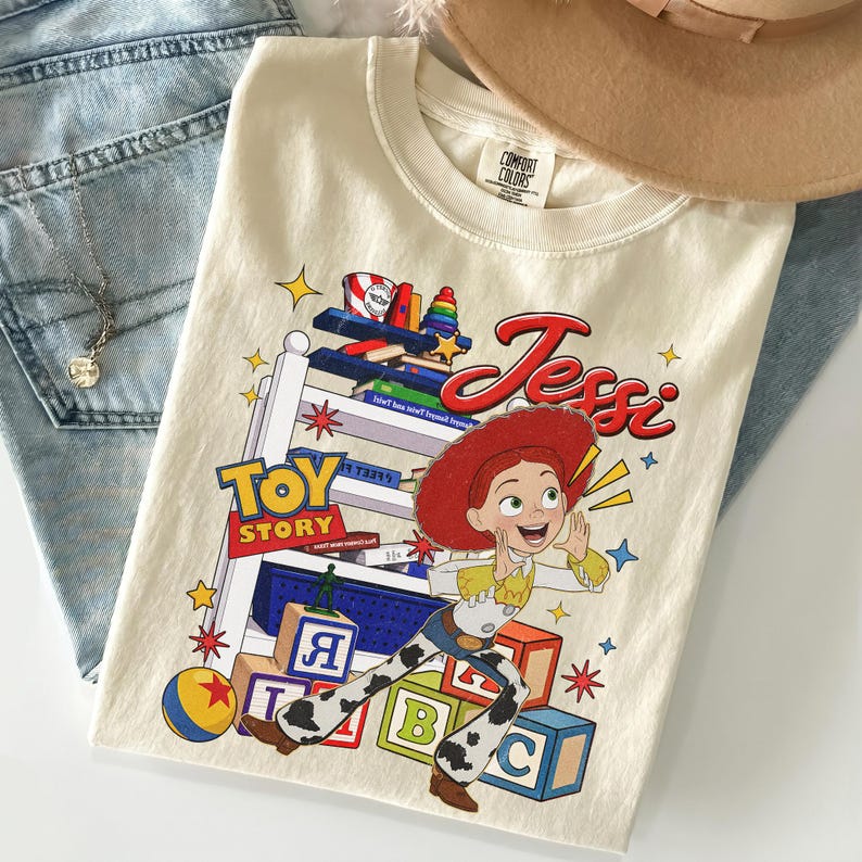 Puede incluir: Camiseta color crema con el personaje Jessi de Toy Story. El dise&ntilde;o incluye las palabras "Toy Story" y "Jessi" en rojo, junto con bloques de juguetes coloridos y estrellas. La camiseta est&aacute; doblada sobre unos vaqueros azules.