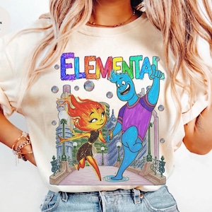 Puede incluir: Camiseta color marfil con la palabra "ELEMENTAL" en colores arcoíris. El gráfico muestra dos personajes de dibujos animados, uno con pelo de fuego y el otro azul, tomados de la mano en un puente.