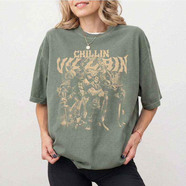 May include: An olive green short-sleeve t-shirt featuring a tan graphic print of various animated villain characters. The text "CHILLIN LIKE A VILLAIN" is printed in a distressed, stylized font above the group of characters on the front of the shirt.