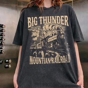 May include: A charcoal grey short-sleeve t-shirt featuring a beige graphic print of a steam locomotive train. The text reads BIG THUNDER FRONTIERLAND MOUNTIAN RAILROAD. The design includes a mountain landscape background with smoke rising from the engine.