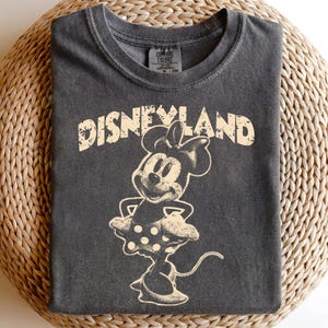 May include: Dark gray t-shirt featuring a distressed graphic of Minnie Mouse and the word "DISNEYLAND" in a vintage style. The shirt has a crew neck and short sleeves. The graphic is in a cream color.