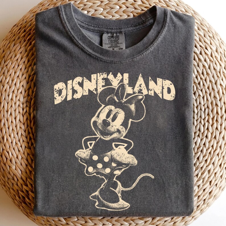 May include: Dark gray t-shirt featuring a distressed graphic of Minnie Mouse and the word "DISNEYLAND" in a vintage style. The shirt has a crew neck and short sleeves. The graphic is in a cream color.