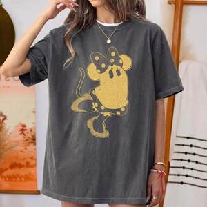 Pode incluir: Camiseta cinza escuro com um gr&aacute;fico dourado da Minnie Mouse. A camiseta oversized tem mangas curtas e gola redonda. O design da Minnie Mouse inclui um la&ccedil;o e um vestido de bolinhas. Um colar dourado &eacute; vis&iacute;vel.