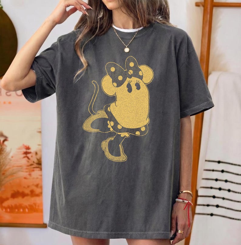 Pode incluir: Camiseta cinza escuro com um gr&aacute;fico dourado da Minnie Mouse. A camiseta oversized tem mangas curtas e gola redonda. O design da Minnie Mouse inclui um la&ccedil;o e um vestido de bolinhas. Um colar dourado &eacute; vis&iacute;vel.