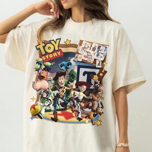 Op de afbeelding: Een crèmekleurig t-shirt met een kleurrijke grafische print van Toy Story-personages, waaronder Woody, Buzz Lightyear, Jessie, Rex en Mr. Potato Head. Het ontwerp bevat het Toy Story-logo en een prikbord met de tekst Andy's Room Keep Out.