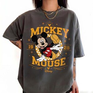May include: Dark gray t-shirt featuring Mickey Mouse. The design includes the words "Mickey Mouse" in yellow, along with the year "1928" and a graphic of Mickey Mouse in a playful pose. The Disney logo is at the bottom.