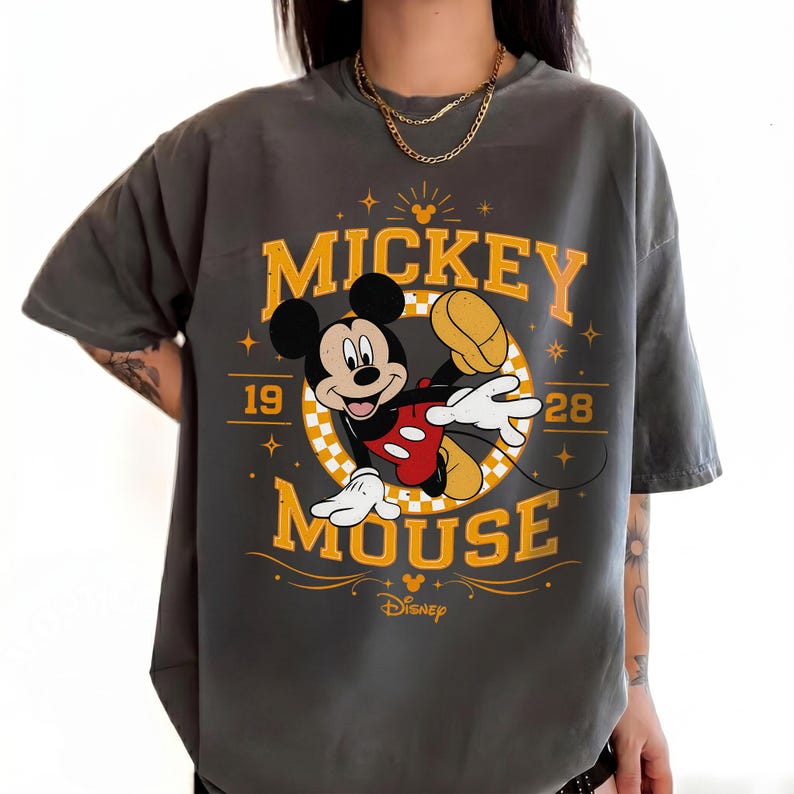 May include: Dark gray t-shirt featuring Mickey Mouse. The design includes the words "Mickey Mouse" in yellow, along with the year "1928" and a graphic of Mickey Mouse in a playful pose. The Disney logo is at the bottom.