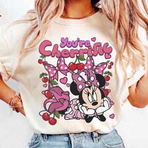 Pode incluir: Camiseta cor marfim com um desenho da Minnie Mouse e o texto "You're Cherrific". O design inclui la&ccedil;os rosa, cerejas e cora&ccedil;&otilde;es. &Eacute; uma pe&ccedil;a de roupa casual.