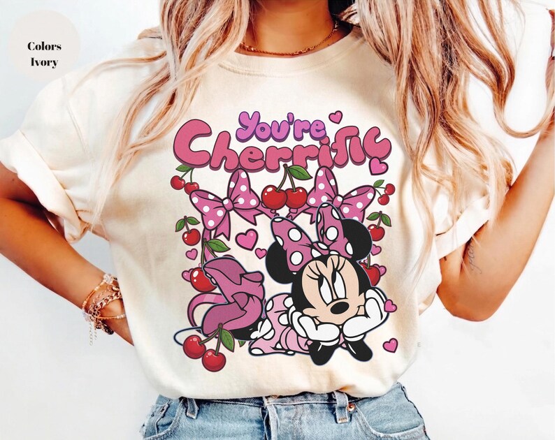 Pode incluir: Camiseta cor marfim com um desenho da Minnie Mouse e o texto "You're Cherrific". O design inclui la&ccedil;os rosa, cerejas e cora&ccedil;&otilde;es. &Eacute; uma pe&ccedil;a de roupa casual.