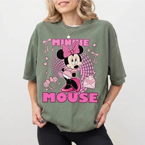 May include: Sage green t-shirt with a Minnie Mouse graphic in pink and black. The design includes the words "MINNIE MOUSE" in pink block letters, along with hearts, bows, and a pink envelope.