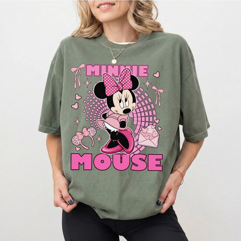 May include: Sage green t-shirt with a Minnie Mouse graphic in pink and black. The design includes the words "MINNIE MOUSE" in pink block letters, along with hearts, bows, and a pink envelope.