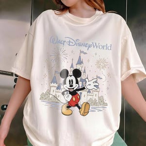 May include: Cream-colored t-shirt featuring a graphic of Mickey Mouse in front of a castle with the text "Walt Disney World." The design includes fireworks and stars. The shirt has short sleeves and a relaxed fit.