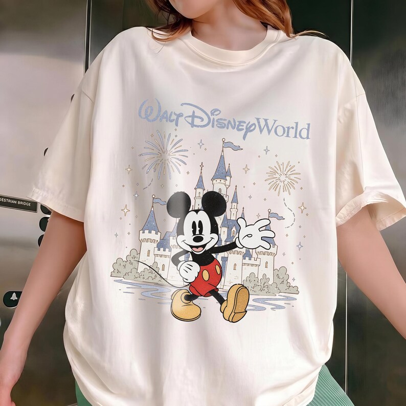 May include: Cream-colored t-shirt featuring a graphic of Mickey Mouse in front of a castle with the text "Walt Disney World." The design includes fireworks and stars. The shirt has short sleeves and a relaxed fit.