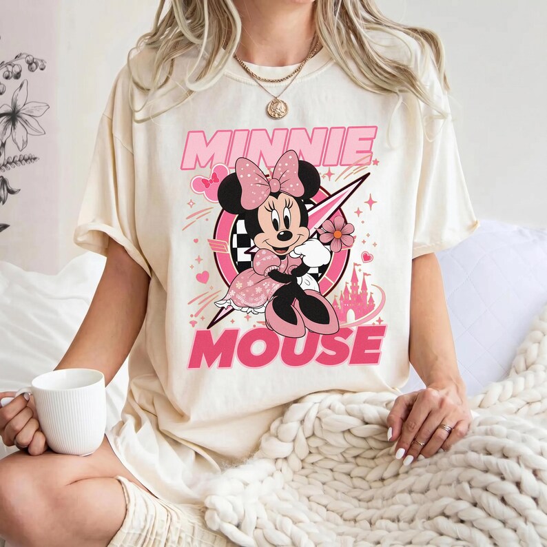 May include: Cream-colored t-shirt featuring a graphic of Minnie Mouse in a pink dress and bow, holding a flower. The design includes the words "MINNIE MOUSE" in pink, with a castle and racing-themed elements.