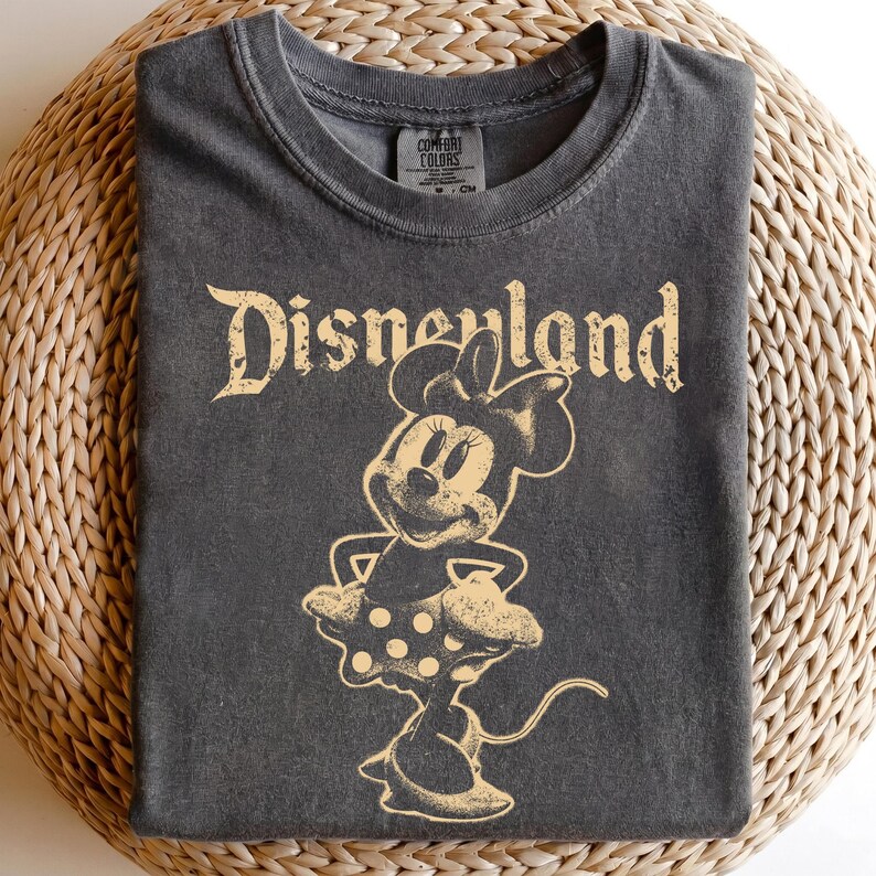 K&ouml;nnte beinhalten: Dunkelgraues T-Shirt mit einem Minnie Mouse-Motiv im Vintage-Stil und dem Wort "Disneyland" in einer verblassten Schriftart. Das Shirt besteht aus einem weichen Material und hat einen Rundhalsausschnitt. Das Shirt liegt auf einer gewebten Oberfl&auml;che.