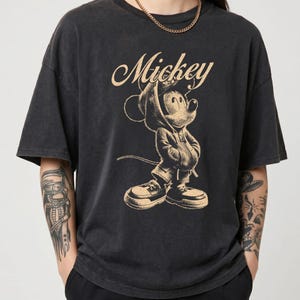 Puede incluir: Una camiseta de manga corta color negro carb&oacute;n con un estampado gr&aacute;fico en beige de Mickey Mouse vistiendo una sudadera con capucha y zapatillas. La palabra Mickey est&aacute; impresa en letra cursiva sobre la ilustraci&oacute;n en la parte frontal.