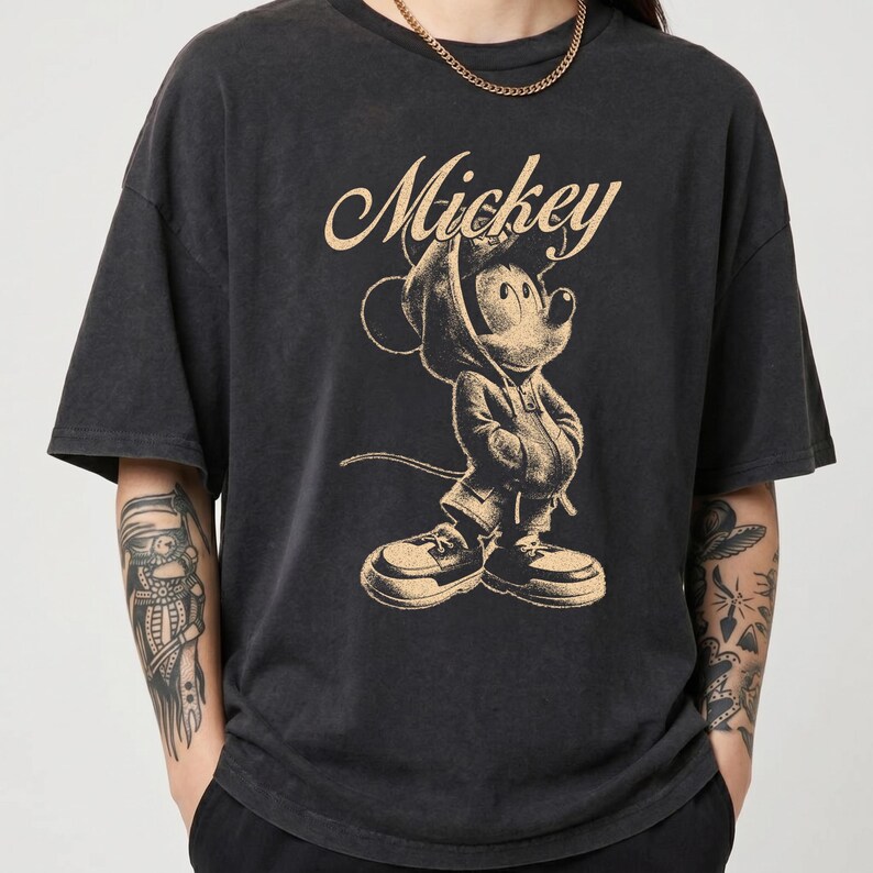 Puede incluir: Una camiseta de manga corta color negro carb&oacute;n con un estampado gr&aacute;fico en beige de Mickey Mouse vistiendo una sudadera con capucha y zapatillas. La palabra Mickey est&aacute; impresa en letra cursiva sobre la ilustraci&oacute;n en la parte frontal.