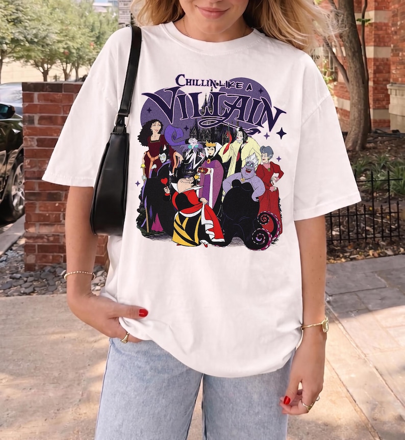 K&ouml;nnte beinhalten: Ein wei&szlig;es T-Shirt mit kurzen &Auml;rmeln und einem Grafikdruck verschiedener animierter B&ouml;sewichte. Der Text "CHILLIN LIKE A VILLAIN" ist in Lila &uuml;ber den Figuren gedruckt. Das Design zeigt Charaktere in roten, schwarzen, violetten und gelben Outfits.