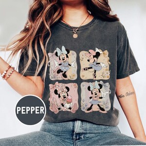 May include: Dark gray t-shirt featuring a four-panel design of Minnie Mouse in various poses. Each panel has a different pastel background with floral accents. Minnie is wearing different outfits and bows in each panel. The word "PEPPER" is printed on a circular label.