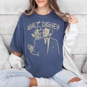 May include: A blue t-shirt featuring a vintage-style graphic of Walt Disney with Mickey Mouse. The text "WALT DISNEY" is arched above the image. The shirt has short sleeves and a relaxed fit.