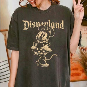 K&ouml;nnte beinhalten: Dunkelgraues T-Shirt mit dem Text "Disneyland" in einer Vintage-Schriftart &uuml;ber einer Minnie Mouse-Grafik. Die Minnie Mouse-Illustration ist im klassischen Stil gehalten und tr&auml;gt einen gepunkteten Rock.