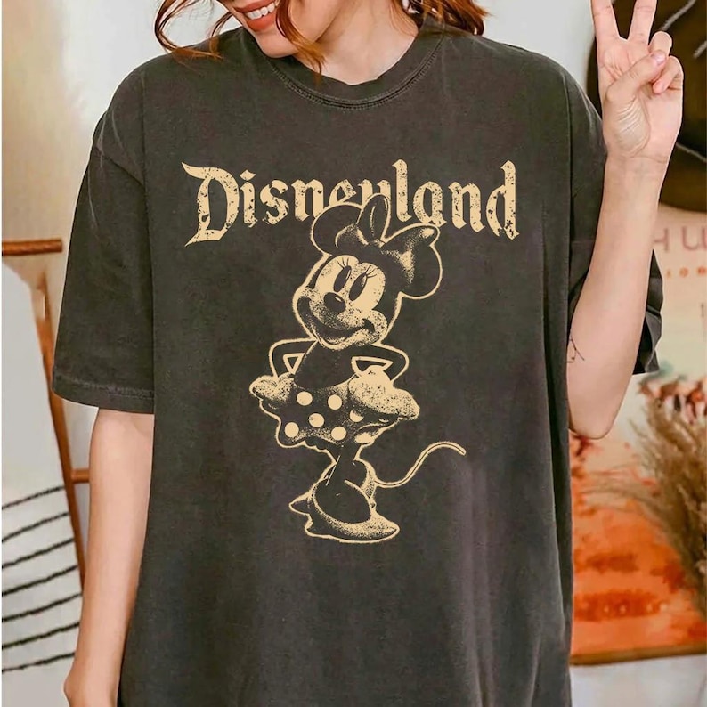 K&ouml;nnte beinhalten: Dunkelgraues T-Shirt mit dem Text "Disneyland" in einer Vintage-Schriftart &uuml;ber einer Minnie Mouse-Grafik. Die Minnie Mouse-Illustration ist im klassischen Stil gehalten und tr&auml;gt einen gepunkteten Rock.