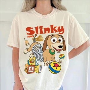 Può includere: T-shirt color crema con un'immagine a cartoni animati di Slinky Dog di Toy Story. Il design include la parola "Slinky" in rosso, l'anno "1995" e blocchi e palline colorate. La maglietta ha maniche corte e girocollo.