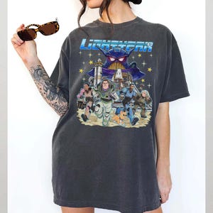 May include: Dark gray t-shirt featuring a colorful graphic from the movie Lightyear. The design includes the movie title, Buzz Lightyear, and other characters running across a sandy landscape. The shirt is a relaxed fit.
