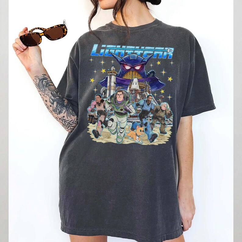 May include: Dark gray t-shirt featuring a colorful graphic from the movie Lightyear. The design includes the movie title, Buzz Lightyear, and other characters running across a sandy landscape. The shirt is a relaxed fit.
