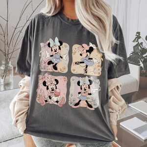 May include: Dark gray t-shirt featuring four squares with Minnie Mouse illustrations. Each square showcases Minnie in different poses and outfits, with floral and bow accents. The shirt has a relaxed fit.