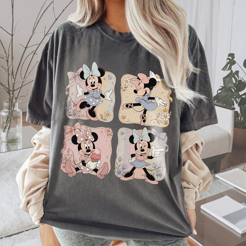 May include: Dark gray t-shirt featuring four squares with Minnie Mouse illustrations. Each square showcases Minnie in different poses and outfits, with floral and bow accents. The shirt has a relaxed fit.