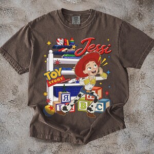 Puede incluir: Camiseta marr&oacute;n con un dise&ntilde;o de Jessi de Toy Story. El dise&ntilde;o incluye las palabras "Toy Story" y "Jessi" en una fuente juguetona, junto con bloques de juguetes coloridos y estrellas. La camiseta tiene un aspecto vintage y desgastado.
