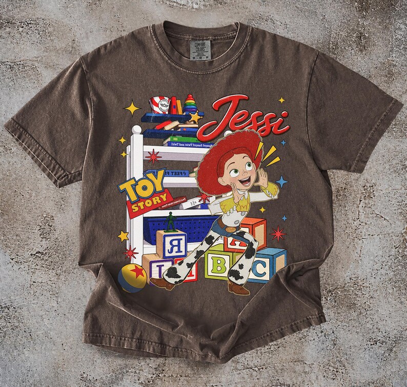 Puede incluir: Camiseta marr&oacute;n con un dise&ntilde;o de Jessi de Toy Story. El dise&ntilde;o incluye las palabras "Toy Story" y "Jessi" en una fuente juguetona, junto con bloques de juguetes coloridos y estrellas. La camiseta tiene un aspecto vintage y desgastado.