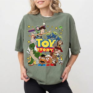 Pu&ograve; includere: T-shirt verde oliva con una grafica colorata dei personaggi del film d'animazione "Toy Story". Il design include Woody, Buzz Lightyear, gli alieni e altri personaggi, con le parole "Toy Story EST 1995."