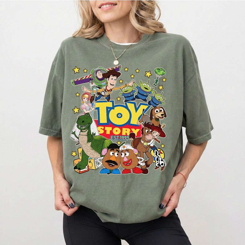 Pu&ograve; includere: T-shirt verde oliva con una grafica colorata dei personaggi del film d'animazione "Toy Story". Il design include Woody, Buzz Lightyear, gli alieni e altri personaggi, con le parole "Toy Story EST 1995."
