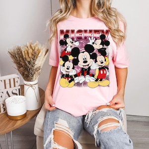 Puede incluir: Camiseta rosa claro con un dise&ntilde;o de Mickey Mouse en varias poses y la palabra "MICKEY" en una fuente retro roja y blanca. La camiseta es de manga corta y tiene cuello redondo.