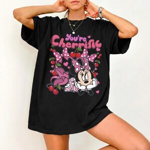 Pode incluir: Camiseta preta com Minnie Mouse com la&ccedil;o e vestido de bolinhas rosa, rodeada de cerejas e la&ccedil;os. O texto "You're Cherrific" est&aacute; escrito em rosa. Um design alegre e divertido.