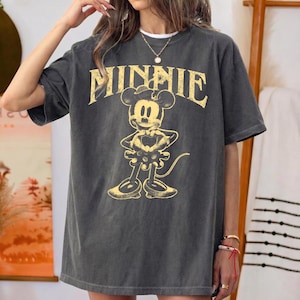 May include: Dark gray t-shirt featuring a distressed yellow graphic of Minnie Mouse with the word "MINNIE" above her. The shirt has short sleeves and a relaxed fit, perfect for casual wear.