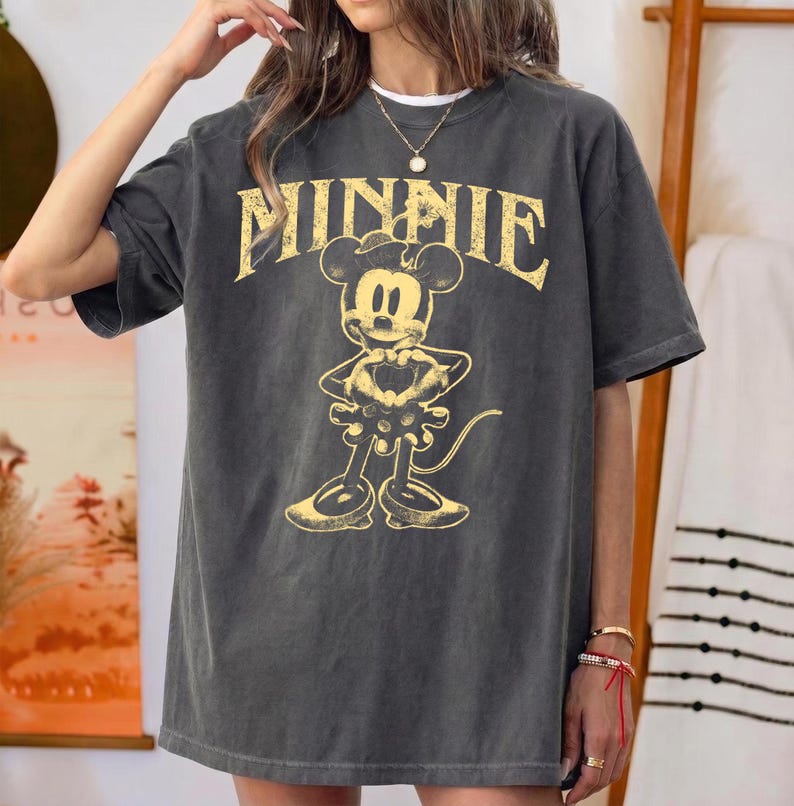 May include: Dark gray t-shirt featuring a distressed yellow graphic of Minnie Mouse with the word "MINNIE" above her. The shirt has short sleeves and a relaxed fit, perfect for casual wear.