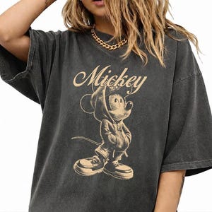 Puede incluir: Una camiseta de manga corta gris carb&oacute;n con un estampado gr&aacute;fico en beige de Mickey Mouse vistiendo una sudadera con capucha y zapatillas. La palabra Mickey est&aacute; impresa en letra cursiva sobre la ilustraci&oacute;n del personaje en el frente.