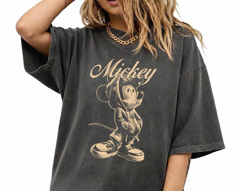 Puede incluir: Una camiseta de manga corta gris carb&oacute;n con un estampado gr&aacute;fico en beige de Mickey Mouse vistiendo una sudadera con capucha y zapatillas. La palabra Mickey est&aacute; impresa en letra cursiva sobre la ilustraci&oacute;n del personaje en el frente.