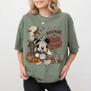 May include: Sage green t-shirt featuring a Halloween-themed graphic. The design includes a haunted house, Mickey Mouse wrapped in bandages, pumpkins, bats, and the text "Nightmare on Main Street."