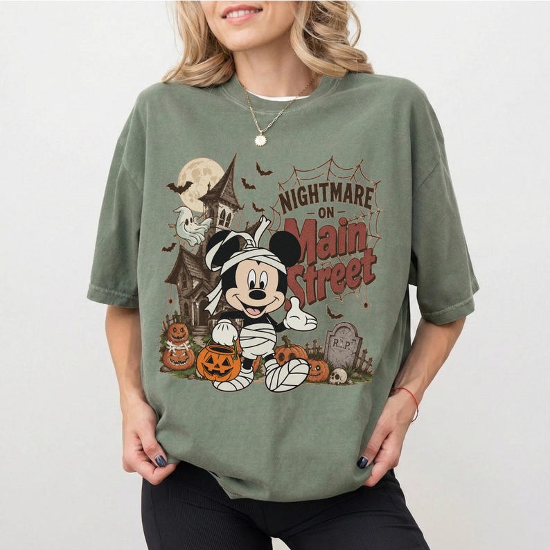 May include: Sage green t-shirt featuring a Halloween-themed graphic. The design includes a haunted house, Mickey Mouse wrapped in bandages, pumpkins, bats, and the text "Nightmare on Main Street."