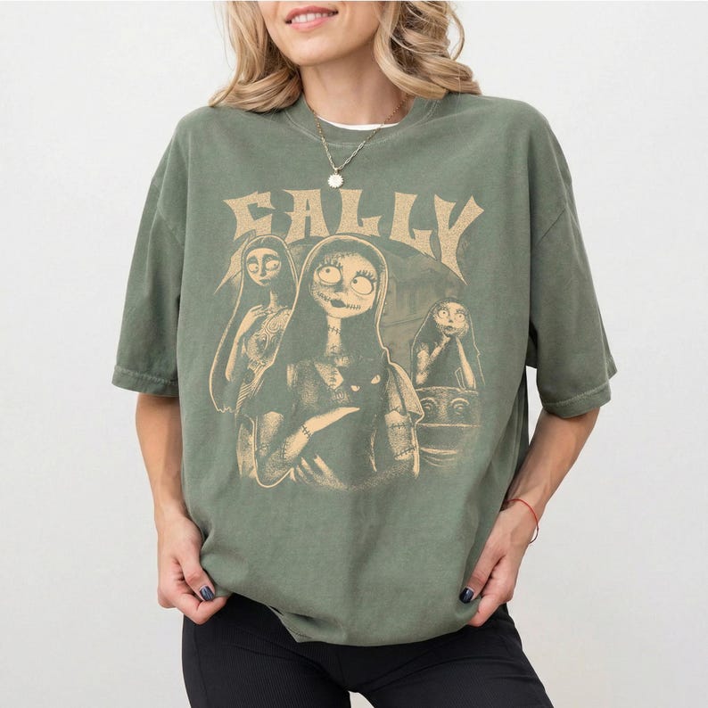 May include: An olive green short-sleeve t-shirt featuring a beige graphic print of the character Sally from The Nightmare Before Christmas. The design includes the text SALLY in a stylized font above three depictions of the character.