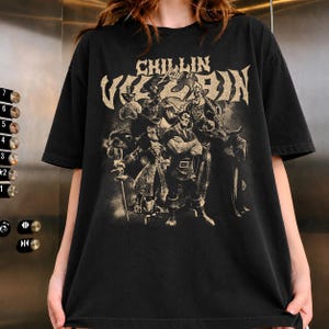 May include: A black short-sleeve t-shirt featuring a beige graphic print of various animated villains. The text "CHILLIN like a VILLAIN" is printed in a stylized font above the characters. The shirt has a crew neckline and a relaxed fit.